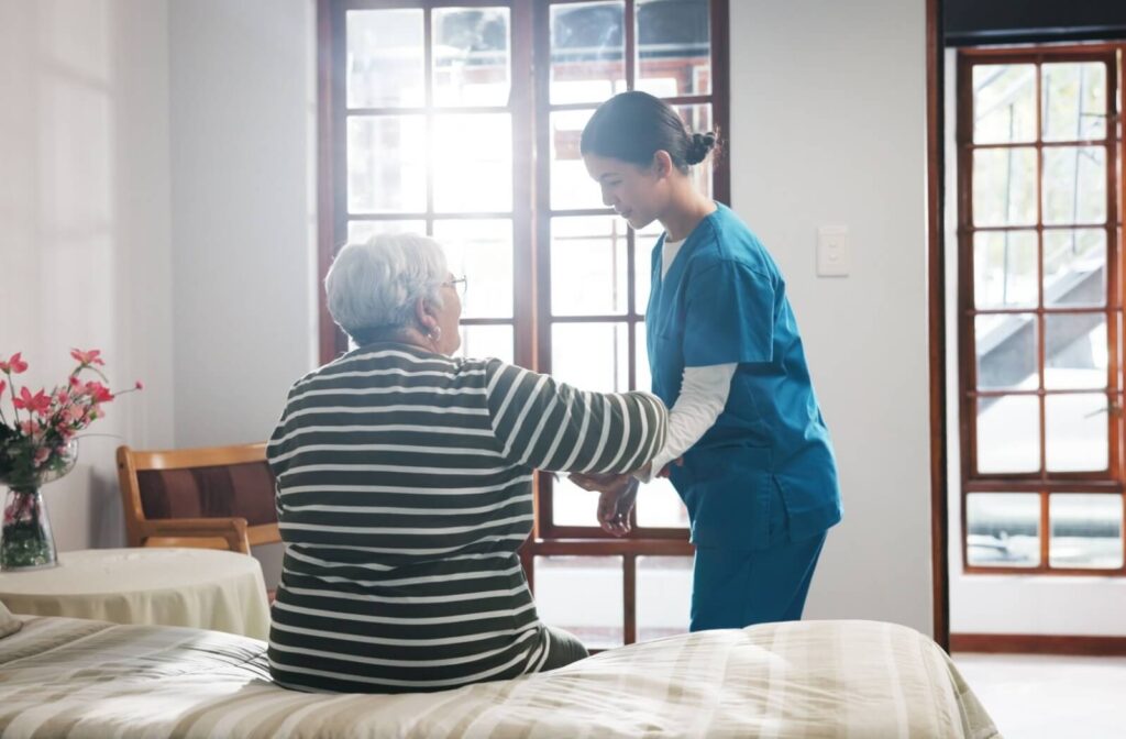 A home healthcare aide gently helps an older adult stand up from their bed in front of a sunlit window